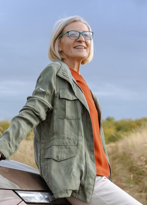 Une femme âgée, adossée à une voiture, regarde au loin avec le sourire, dans un cadre extérieur naturel.