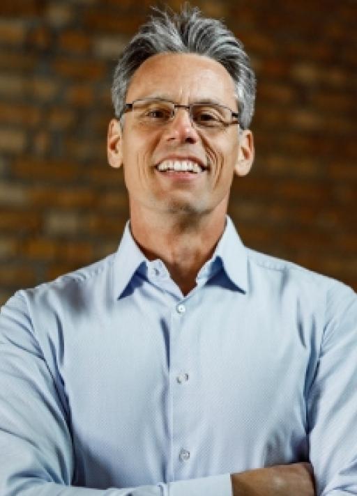 Un homme souriant portant une chemise bleue claire et des lunettes pose devant un mur de briques, les bras croisés.