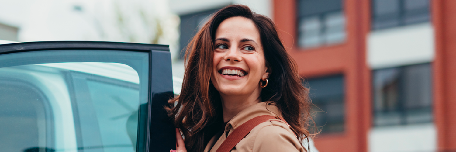 Une femme souriante ouvre la porte d’une voiture blanche en ville, portant un manteau beige et un sac en bandoulière.