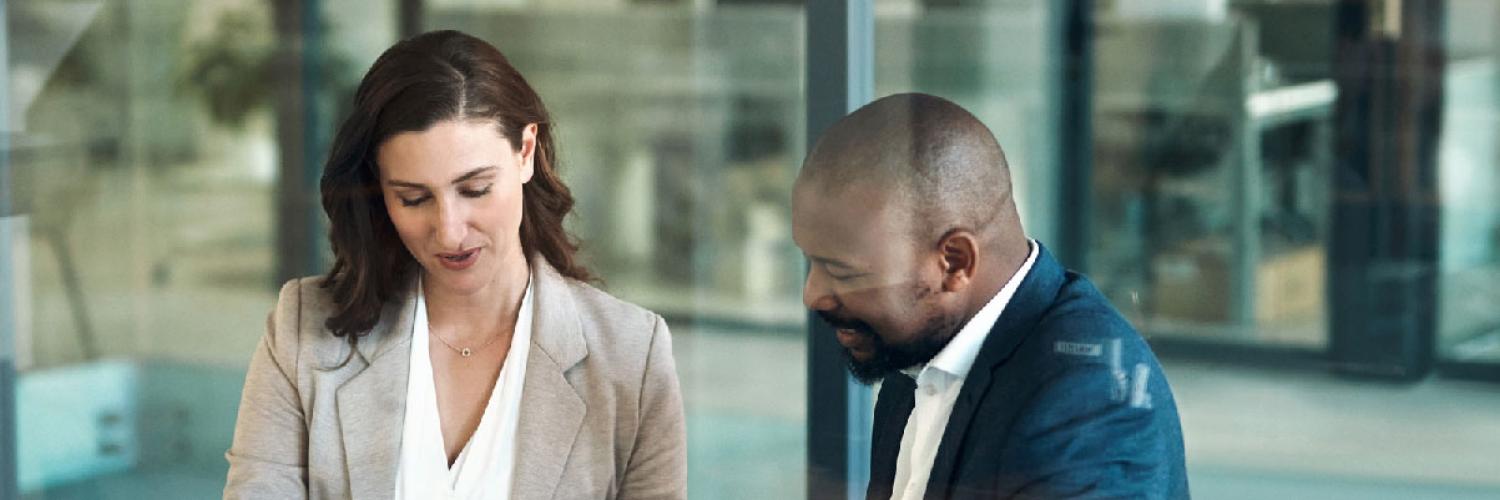 Un homme et une femme en tenue professionnelle discutent et signent des documents à une table de bureau dans un environnement de travail moderne.