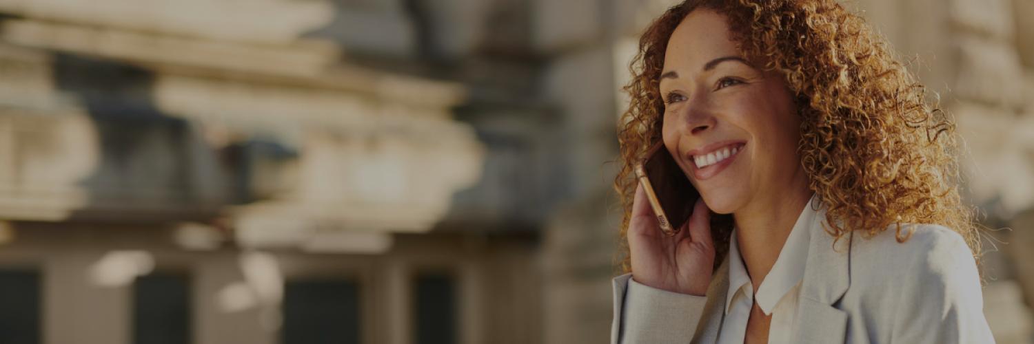 Femme souriante tenant un smartphone lors d'un appel téléphonique devant un bâtiment en ville.